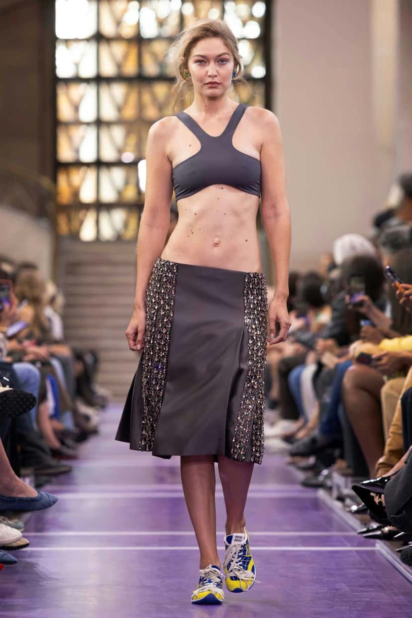 Foto oficial del desfile Primavera/Verano 2027 de Miu Miu: Gigi Hadid en la pasarela con top deportivo gris, pollera gris con paneles bordados y New Balance RC150 azules y amarillas