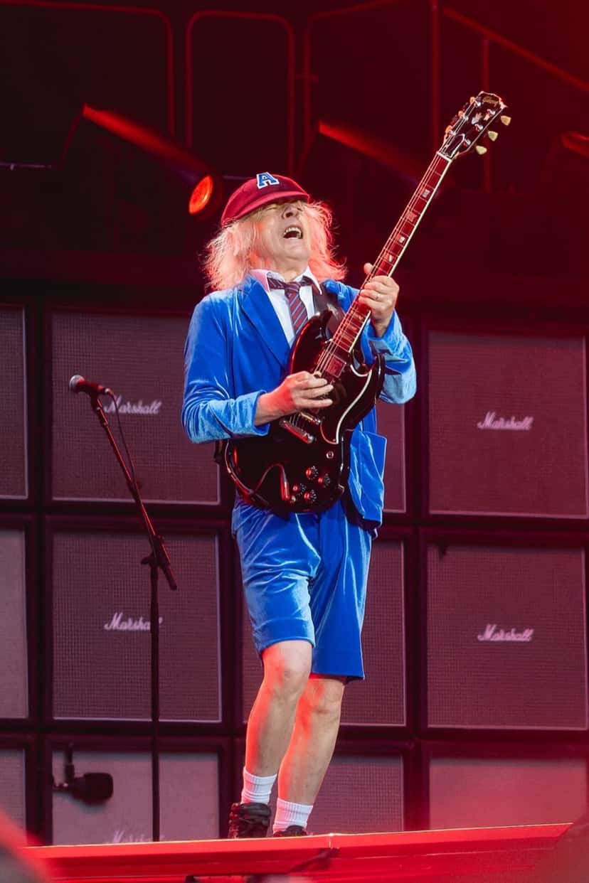 Foto de archivo de Angus Young con gorra bordó, saco y bermudas de terciopelo azul y su guitarra Gibson, tocando frente a una pared de amplificadores Marshall durante la gira Power Up en el estadio de Wembley, en julio de 2024. Foto: Raph_PH, Wikimedia Commons, CC BY 2.0