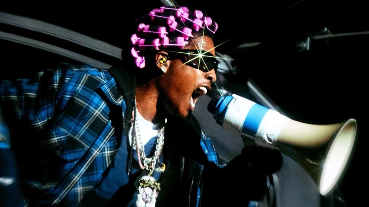 Imagen oficial de la gira Don't Be Dumb World Tour: A$AP Rocky, con ruleros rosas, anteojos negros, camisa escocesa azul y cadenas plateadas, grita por un megáfono azul y blanco asomado a la ventanilla de un auto, sobre un fondo negro