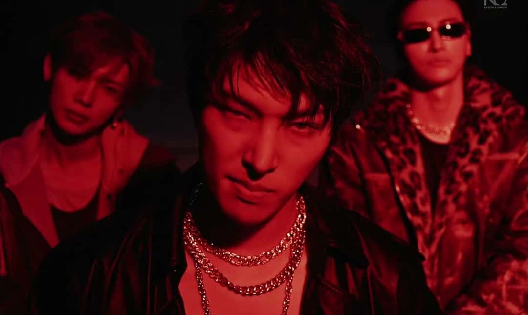 Fotograma del video oficial de «Drip»: tres integrantes de ATEEZ bañados de luz roja, uno adelante con cadenas plateadas y dos atrás, uno de ellos con anteojos de sol y campera de leopardo