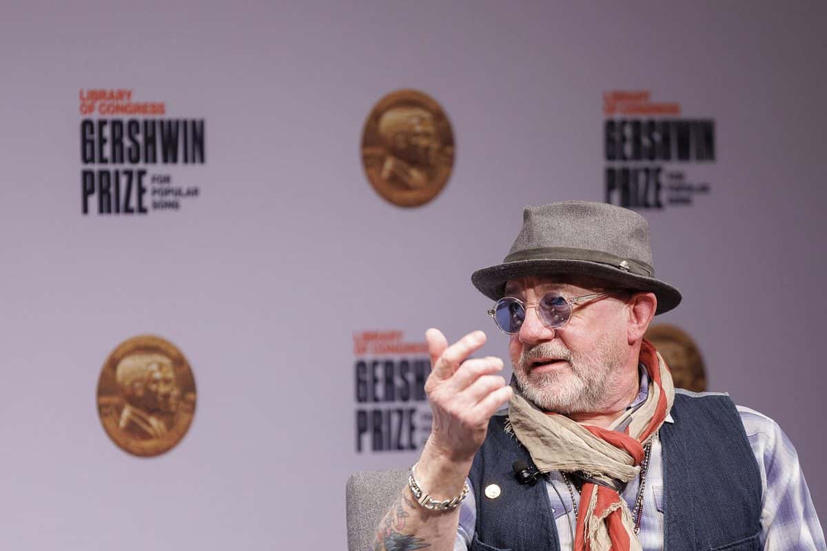 Bernie Taupin con sombrero gris, anteojos redondos y bufanda de colores, hablando y gesticulando con la mano frente a un fondo del Premio Gershwin en la Biblioteca del Congreso de Estados Unidos, en 2024 (foto de archivo). Foto: Shawn Miller/Library of Congress, CC0, vía Wikimedia Commons