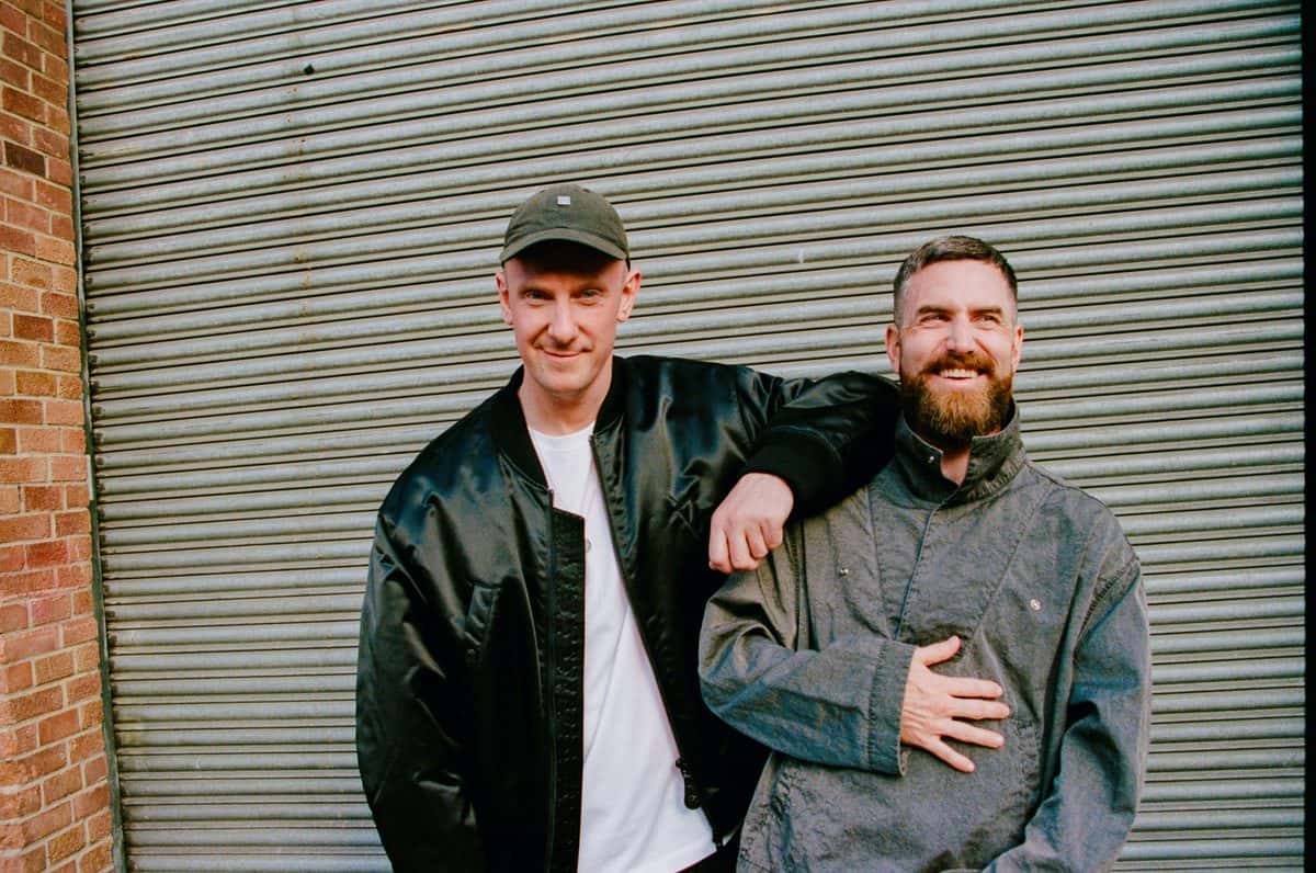 Foto de prensa de Bicep: los dos integrantes del dúo sonriendo frente a una persiana metálica gris, uno con gorra, campera negra y remera blanca y el otro con barba y chaqueta gris