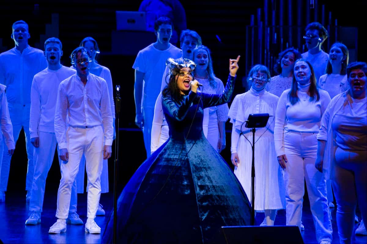Foto oficial de Björk en el concierto Orkestral del 24 de octubre de 2021 en el Harpa de Reikiavik: con un vestido de terciopelo azul y un tocado metálico, canta con el brazo en alto delante del coro Hamrahlíð, todo vestido de blanco