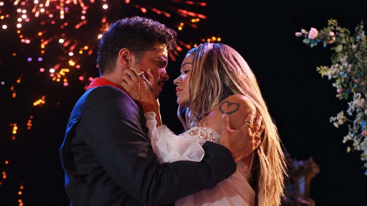 Bruno Mars y Karol G abrazados en una escena del video de Dance With Me, con fuegos artificiales de fondo