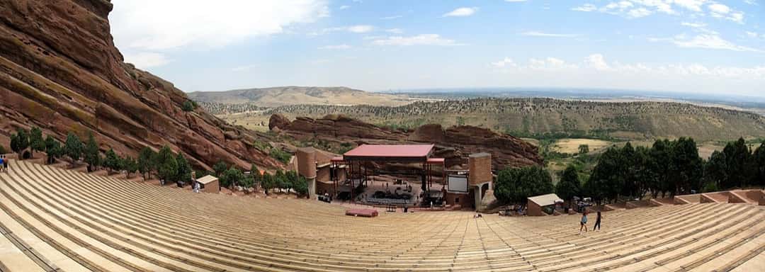 Vista panorámica del Red Rocks Amphitheatre, en Morrison, Colorado: las gradas de piedra bajan entre enormes formaciones de roca rojiza hacia un escenario techado, con el valle y el cielo despejado de fondo. Foto: Jesse Goodier, Wikimedia Commons, dominio público