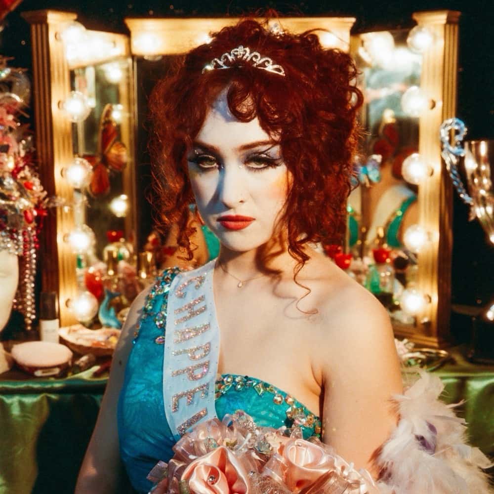 Portada de «The Rise and Fall of a Midwest Princess», de Chappell Roan: la cantante, de pelo colorado y vestido celeste con banda de reina, posa frente a un espejo de camarín con luces