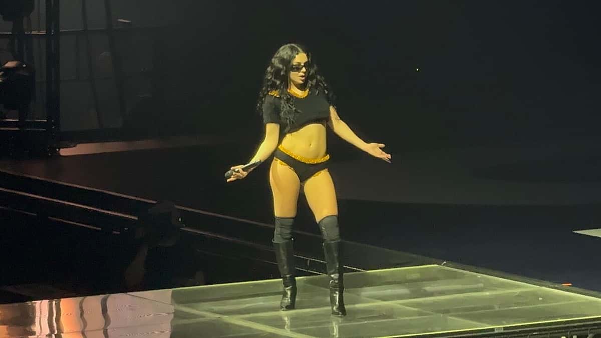 Foto de archivo de Charli XCX en el escenario en abril de 2025: con anteojos de sol, top negro corto, bombacha amarilla y botas negras altas, abre los brazos con el micrófono en la mano sobre una plataforma iluminada. Foto: Erin Mc, Wikimedia Commons, CC BY 2.0