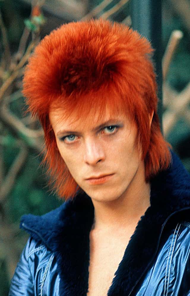 Foto de archivo de David Bowie en los años 70, con el pelo rojo y en punta y una campera azul brillante con cuello de corderito, mirando a cámara. Imagen publicada en el sitio oficial de Bowie80