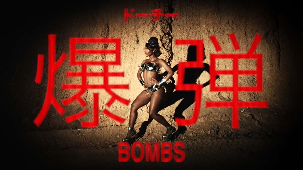 Imagen oficial del video de "Bombs": Doechii, con un conjunto metálico de dos piezas y plataformas, posa con las manos en la cintura contra una pared de tierra iluminada, entre dos grandes caracteres japoneses en rojo y la palabra BOMBS abajo
