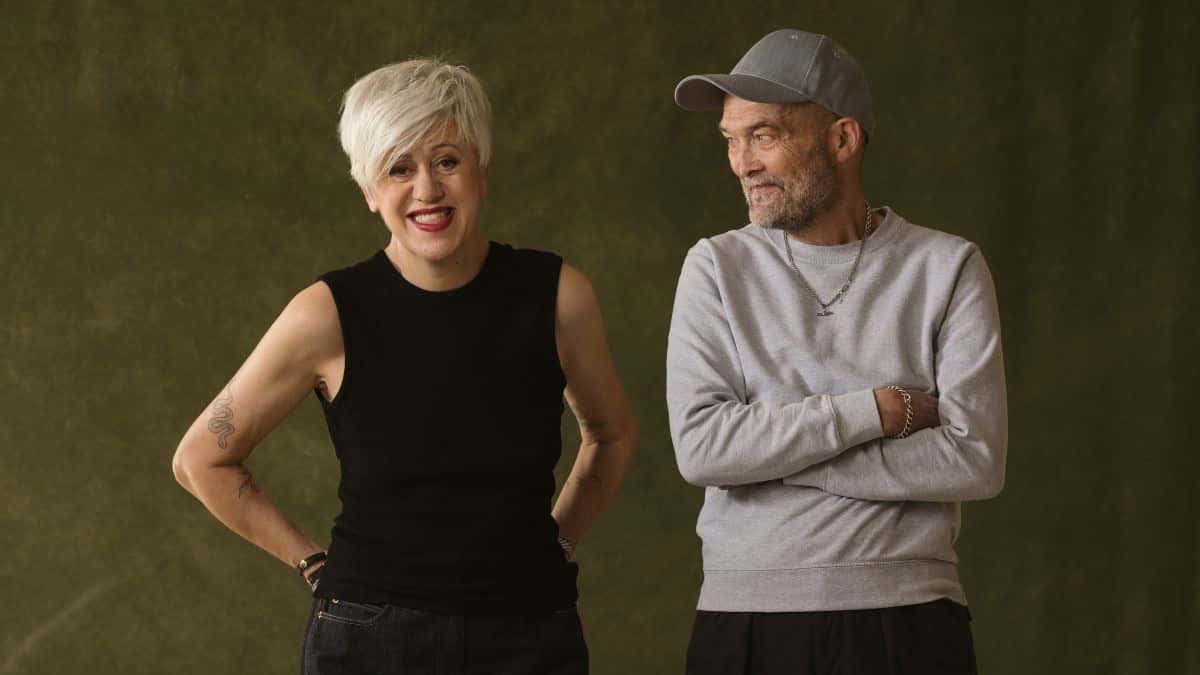 Foto de prensa de Everything But the Girl para «Blue Reservoir»: Tracey Thorn, de pelo corto blanco y musculosa negra, sonríe con las manos en la cintura junto a Ben Watt, con gorra gris y buzo gris de brazos cruzados, sobre un fondo verde oscuro