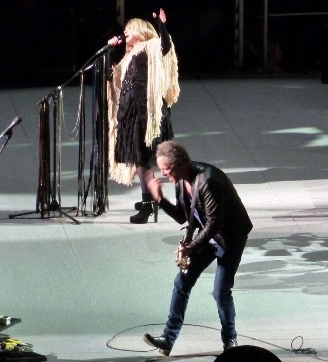 Stevie Nicks y Lindsey Buckingham en vivo en el NIA de Birmingham en noviembre de 2009: ella canta con un chal de flecos claros y el brazo en alto, y él toca la guitarra inclinado hacia adelante, con campera de cuero (foto de archivo). Foto: Tony Hisgett, CC BY 2.0, vía Wikimedia Commons