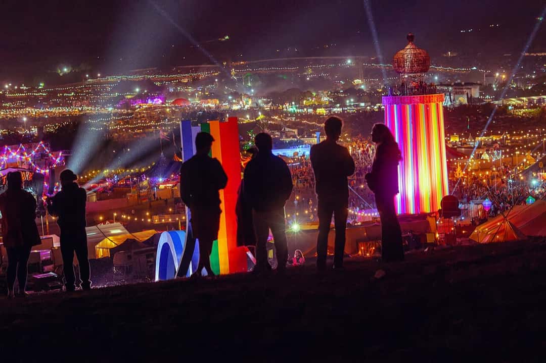 Un grupo de personas a contraluz mira de noche el predio de Glastonbury 2025 desde una colina, con una torre de cintas de colores iluminada, haces de luz y miles de luces de carpas y escenarios de fondo. Foto: Raph_PH, Wikimedia Commons, CC BY 4.0