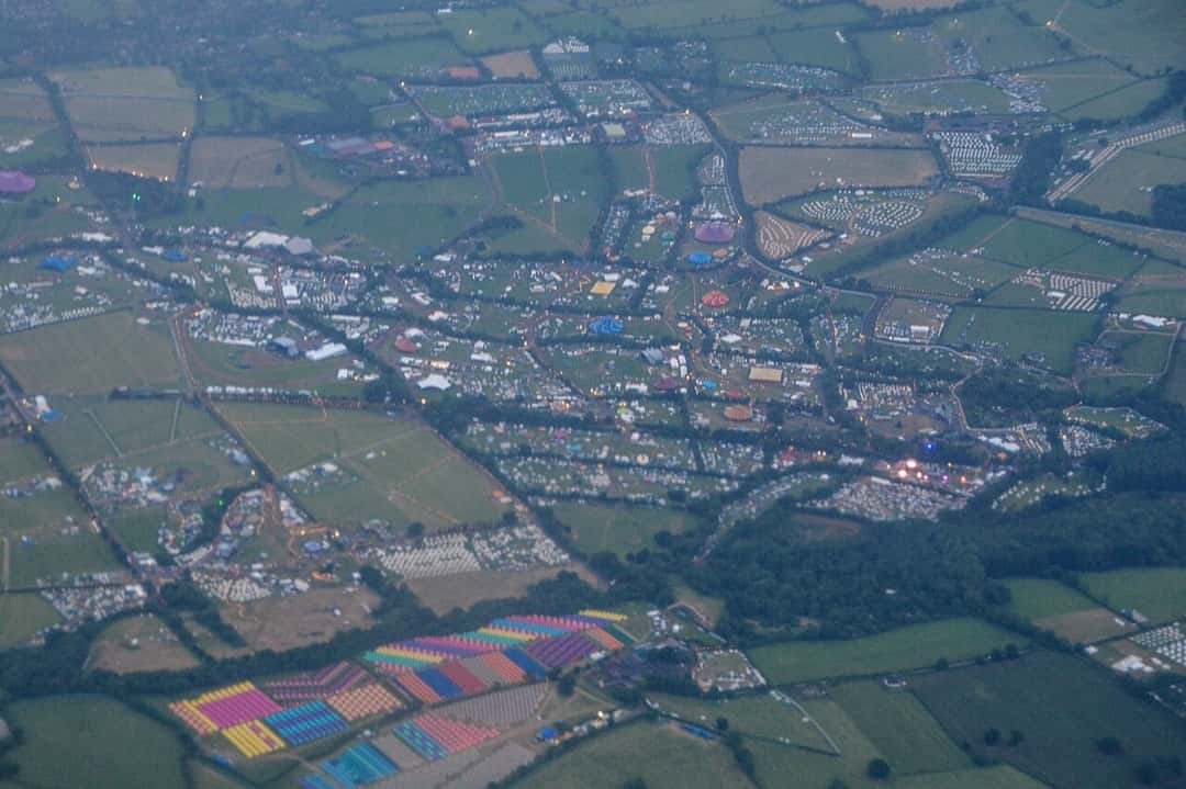 Vista aérea del predio de Glastonbury en Worthy Farm, con campos verdes cubiertos de carpas, carpas de circo de colores y caminos iluminados entre los cercos. Foto: Lewis Clarke, Wikimedia Commons, CC BY-SA 2.0