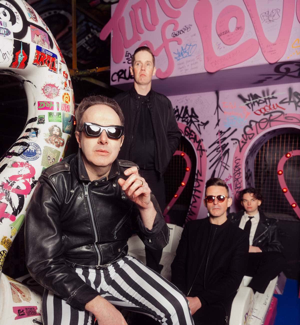 Foto de prensa de Glasvegas: uno de los integrantes, con anteojos negros, campera de cuero y pantalón a rayas, sentado adelante, y el resto de la banda detrás, vestidos de negro, frente a una pared con grafitis