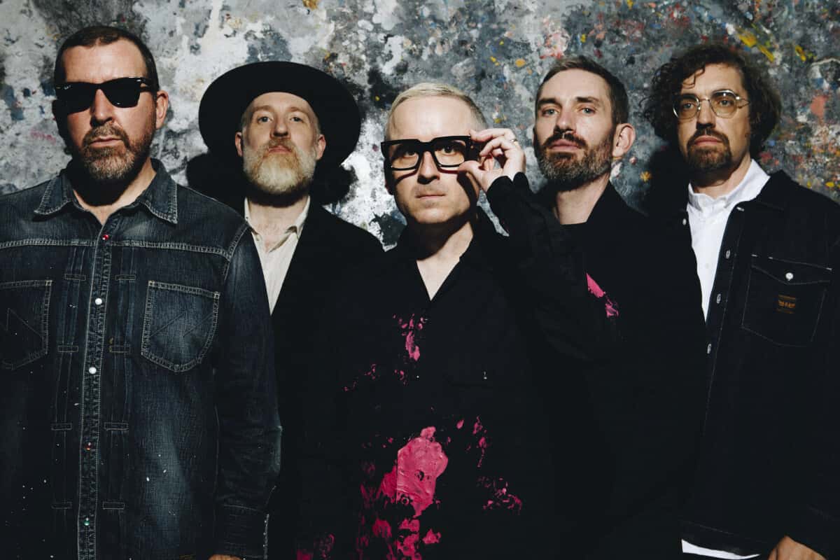 Foto de prensa de Hot Chip: los cinco integrantes de la banda vestidos de negro y de jean, parados frente a una pared manchada de pintura de colores; el del centro se acomoda unos anteojos de marco grueso negro
