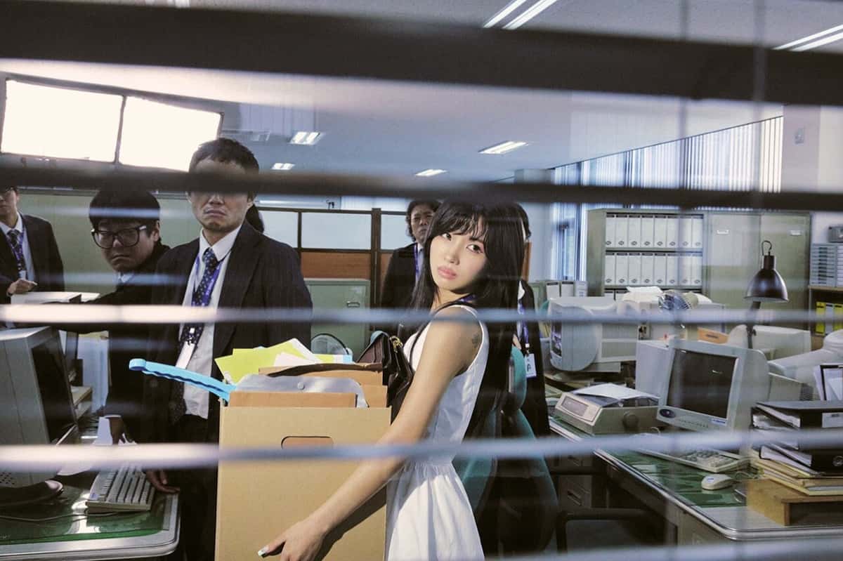 Foto conceptual oficial de Soyeon para "What a Wonderful Life": vestida de blanco, carga una caja de cartón con sus cosas por una oficina llena de monitores viejos mientras sus compañeros de traje la miran, vista a través de una persiana