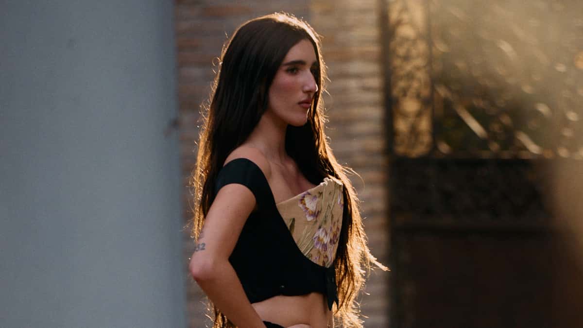 Foto oficial de Judeline para "La Falda": la cantante, de perfil y con el pelo largo iluminado a contraluz, lleva un top negro con un corsé de flores bordadas