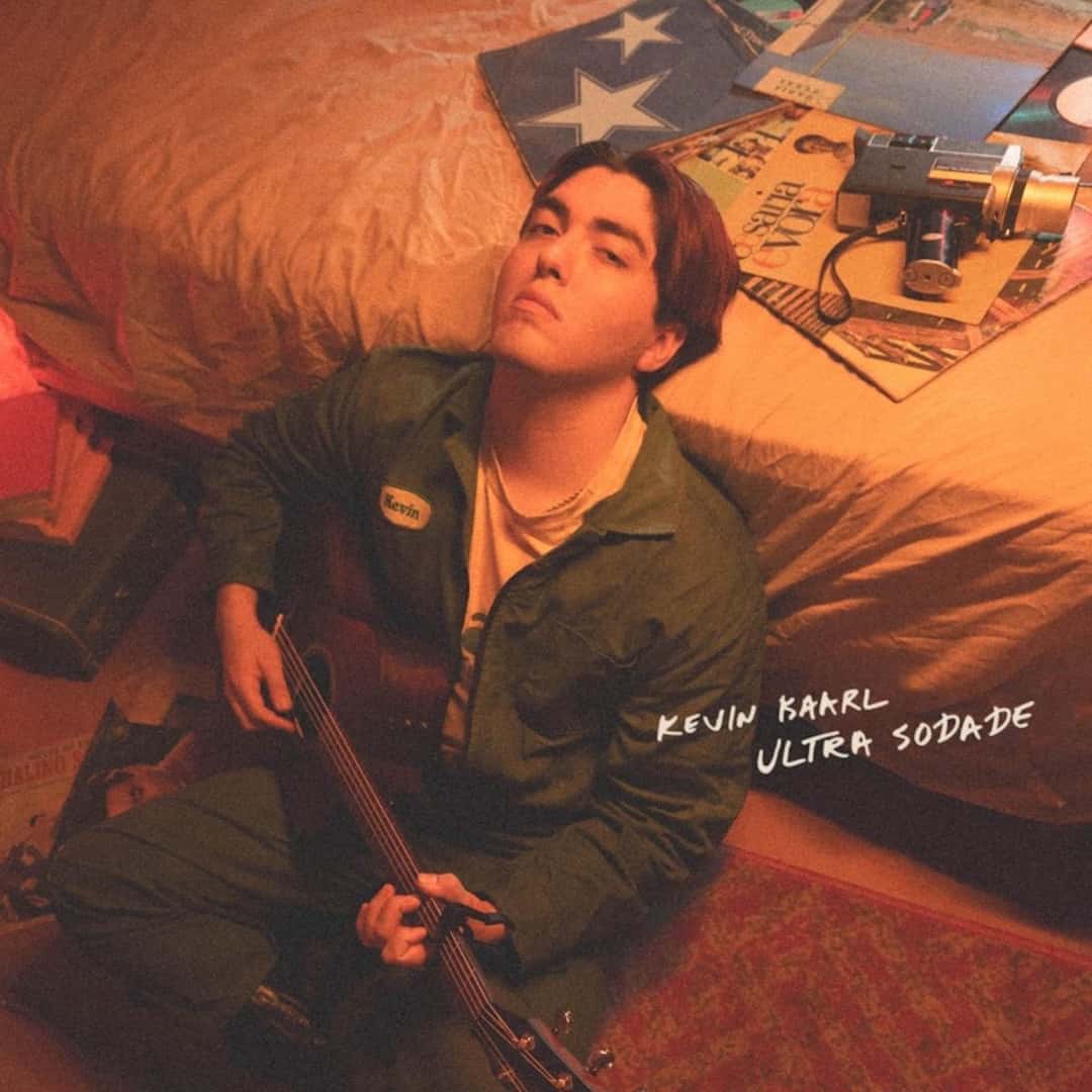 Portada de «ULTRA SODADE», de Kevin Kaarl: el cantante sentado en el piso junto a una cama, con una guitarra, mameluco verde y discos de vinilo detrás