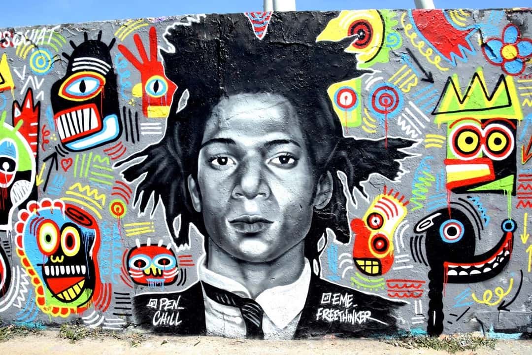 Un mural con el retrato de Jean-Michel Basquiat en blanco y negro, rodeado de figuras coloridas, coronas y caras al estilo de su obra, en el Mauerpark de Berlín, firmado por Eme Freethinker y Pen Chill (imagen que representa al artista). Foto: Singlespeedfahrer, CC0, vía Wikimedia Commons