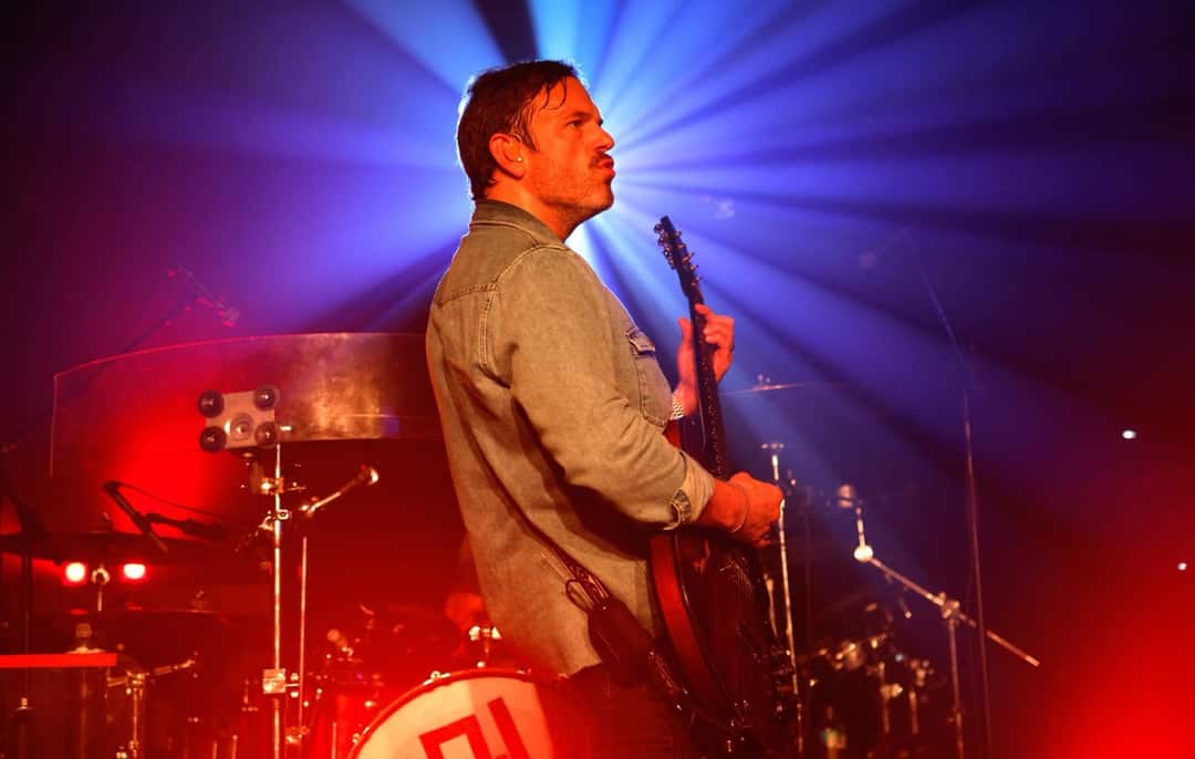 Caleb Followill de perfil tocando la guitarra eléctrica, con camisa de jean, frente a un haz de luz azul y violeta, con una batería iluminada en rojo detrás