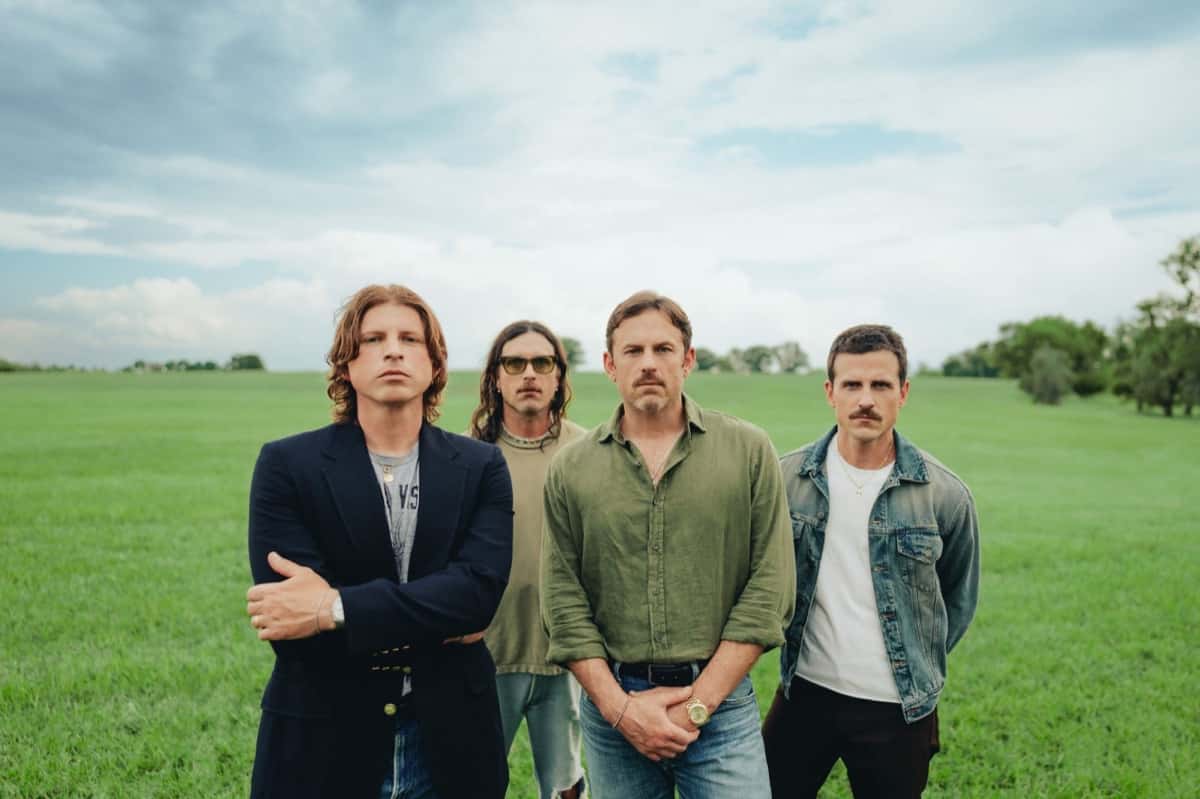 Los cuatro integrantes de Kings of Leon parados en un campo de pasto verde bajo un cielo nublado, uno de saco azul con los brazos cruzados, otro con anteojos amarillos, otro de camisa verde y otro de campera de jean
