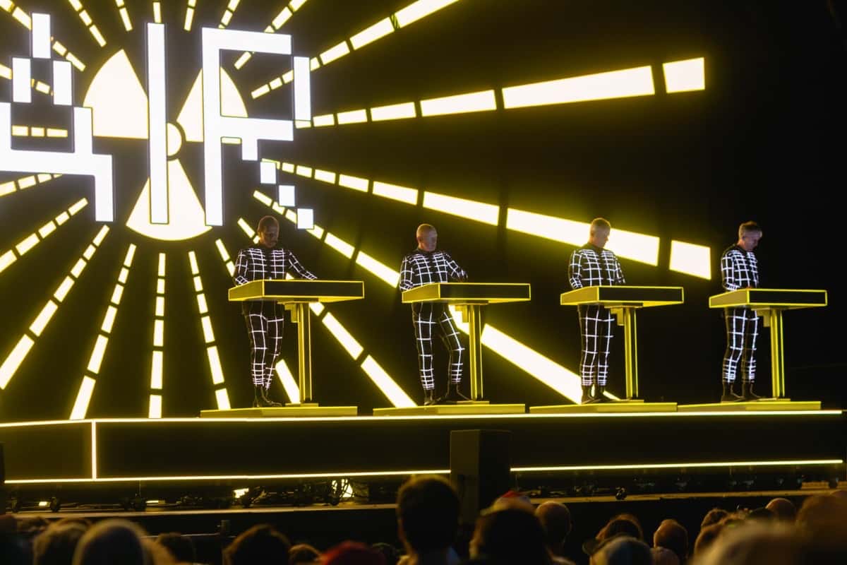 Kraftwerk en vivo en el Forever Now Fest de Milton Keynes en junio de 2025: los cuatro integrantes, con trajes negros cuadriculados con líneas blancas luminosas, detrás de sus consolas amarillas frente a una pantalla con rayos de luz (foto de archivo). Foto: Raph_PH, CC BY 4.0, vía Wikimedia Commons