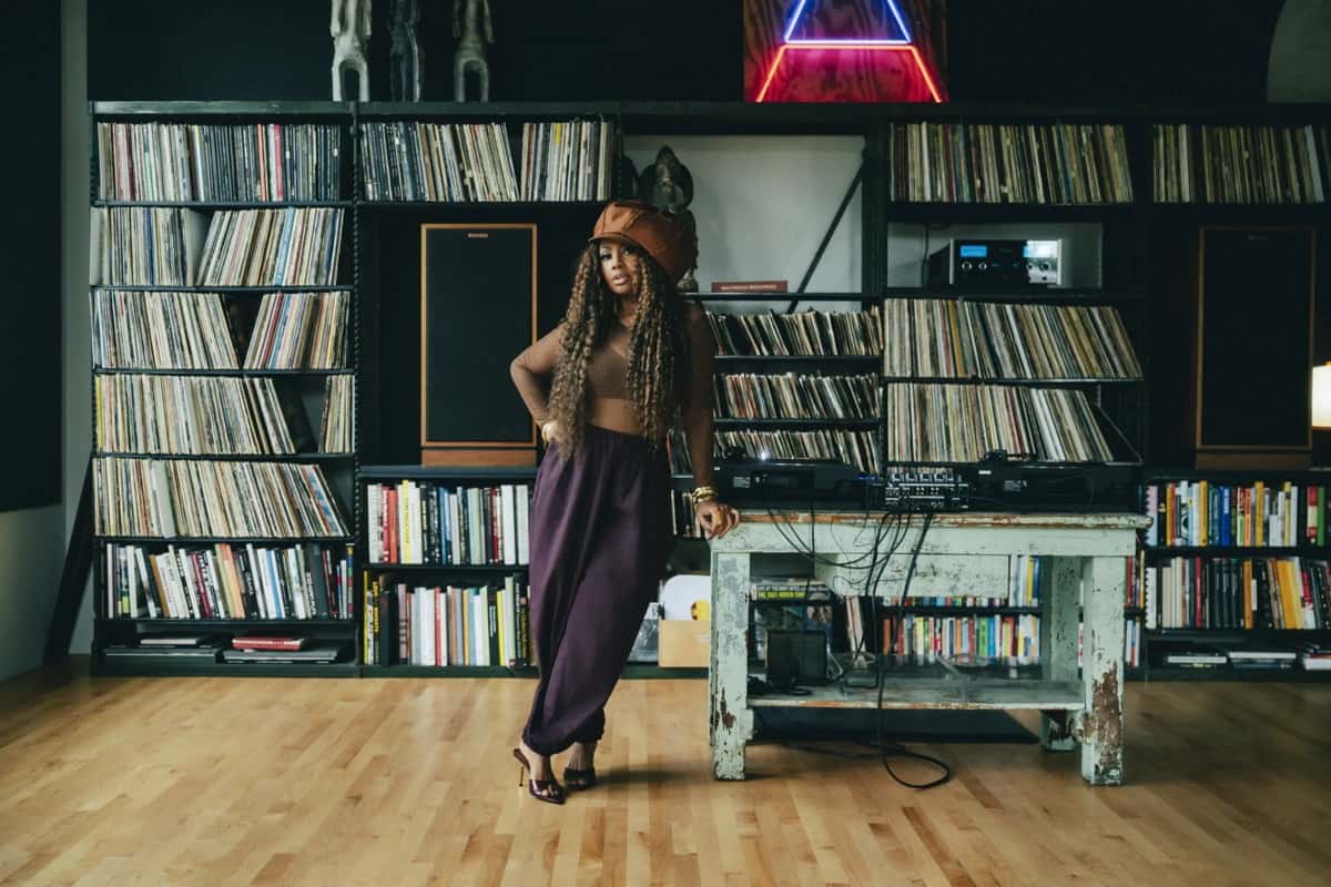 Lalah Hathaway con gorra marrón, top de color tostado y pantalón ancho bordó, apoyada en una mesa de madera gastada con bandejas de DJ, frente a una biblioteca llena de discos de vinilo y libros