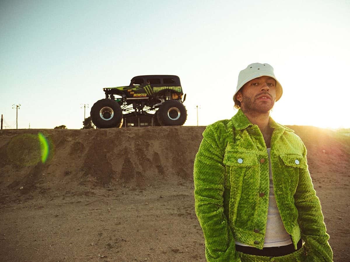 Feid, con piluso blanco y campera de corderoy verde, posa en un descampado de tierra al atardecer, con una camioneta monster truck arriba de una loma, en marzo de 2022 (foto de archivo)