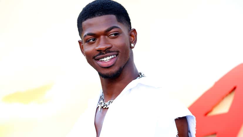 Lil Nas X sonriendo, con dientes con brillantes, camisa blanca sin mangas y cadena plateada, en una alfombra roja con fondo amarillo y rojo