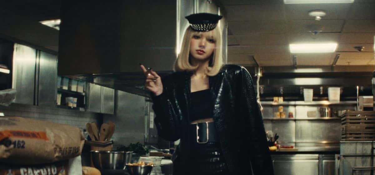 Fotograma del video oficial de «Tarantino»: LISA, con peluca rubia carré, gorra negra de visera y saco de cuero negro, parada en la cocina industrial de un hotel