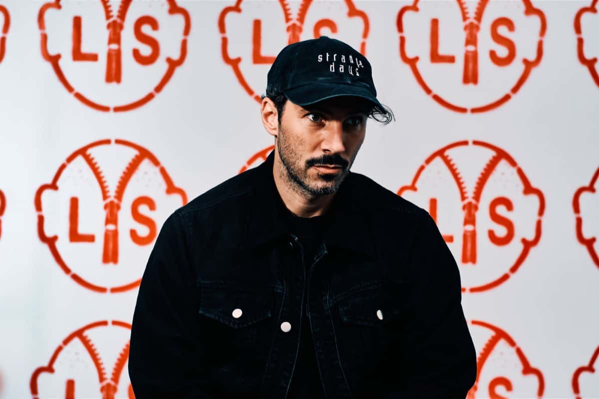 Foto de prensa oficial de Lorenzo Senni para "All Ages", por Jim C. Nedd: el productor, con gorra y campera de jean negras, sentado frente a una pared llena del logo rojo del disco