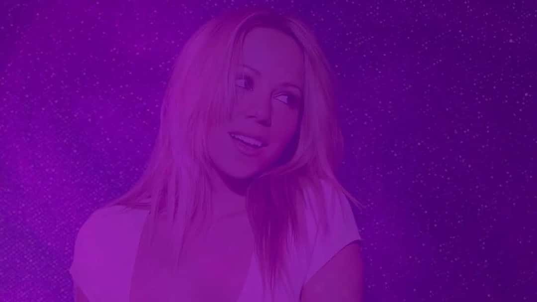 Mariah Carey, de pelo rubio largo, mira de costado y sonríe en una foto de la época de «Glitter» teñida de violeta, en otra imagen del lyric video oficial de «Didn't Mean to Turn You On»