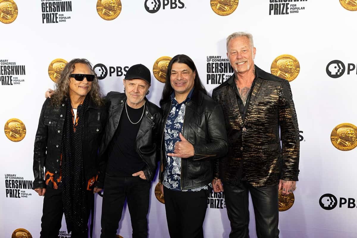 Foto de archivo de Metallica: Kirk Hammett con anteojos negros, Lars Ulrich con gorra, Robert Trujillo con camisa estampada y James Hetfield con saco dorado y negro, posando abrazados frente a un panel blanco del Premio Gershwin en 2024
