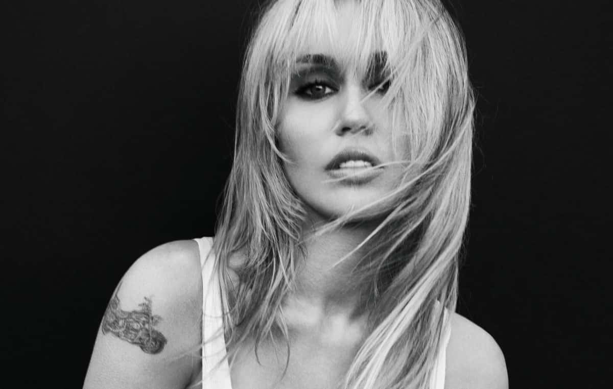Foto de prensa oficial de Miley Cyrus para la etapa de "Bass Persuades", en blanco y negro: con flequillo y el pelo rubio largo cruzándole la cara, musculosa blanca y un tatuaje en el brazo derecho, mira a cámara sobre un fondo negro. Foto: Mert Alas
