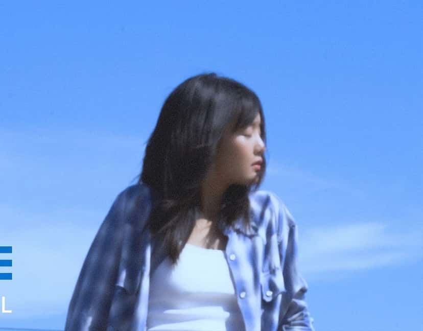 Fotograma de un adelanto oficial del EP: MINNIE, con camisa a cuadros celeste abierta sobre una musculosa blanca, mira hacia un costado con el cielo azul de fondo