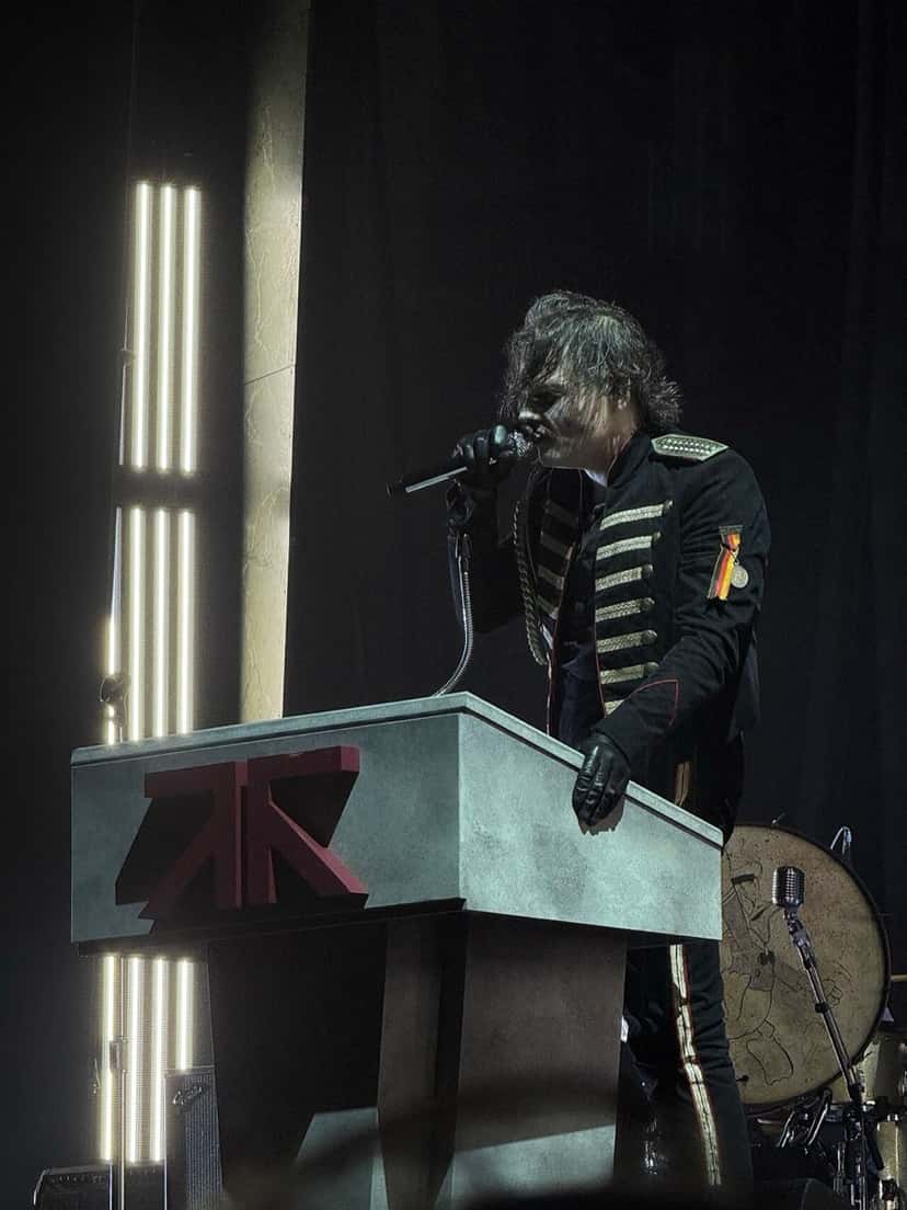 Gerard Way canta detrás de un atril gris con el logo de la banda, con uniforme militar negro, guantes y luces blancas verticales detrás, en un show de la gira de The Black Parade en Tampa (foto de archivo, septiembre de 2025). Foto: Ollip182, dominio público (CC0), vía Wikimedia Commons