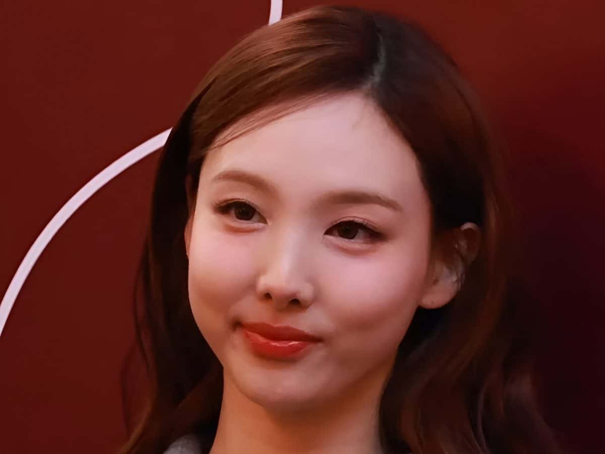 Nayeon de TWICE sonriendo en un evento de Maison Kitsuné en octubre de 2024, con pelo castaño suelto y campera gris con cierre, frente a un fondo rojo oscuro (foto de archivo). Foto: TV10, CC BY 3.0, vía Wikimedia Commons