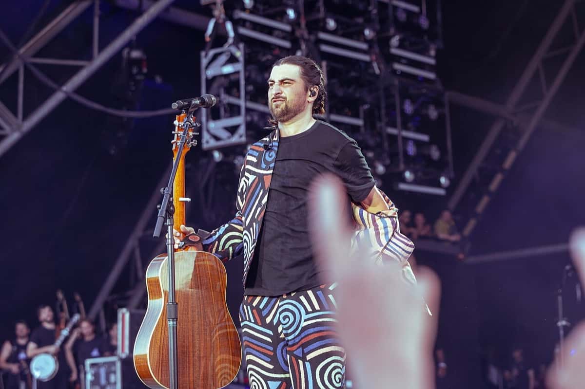 Noah Kahan en el escenario del Glastonbury Festival de 2025, con un traje de estampado de espirales de colores, una remera negra y una guitarra acústica a un costado, frente a una estructura de luces (foto de archivo)