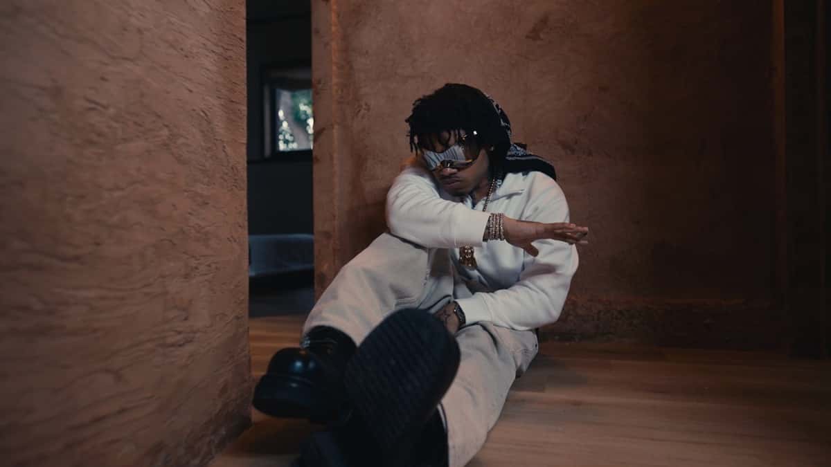 NoCap sentado en el piso de madera de un pasillo de paredes color terracota, con lentes plateados, pañuelo en la cabeza y conjunto claro, en una imagen del video de Angels Smile