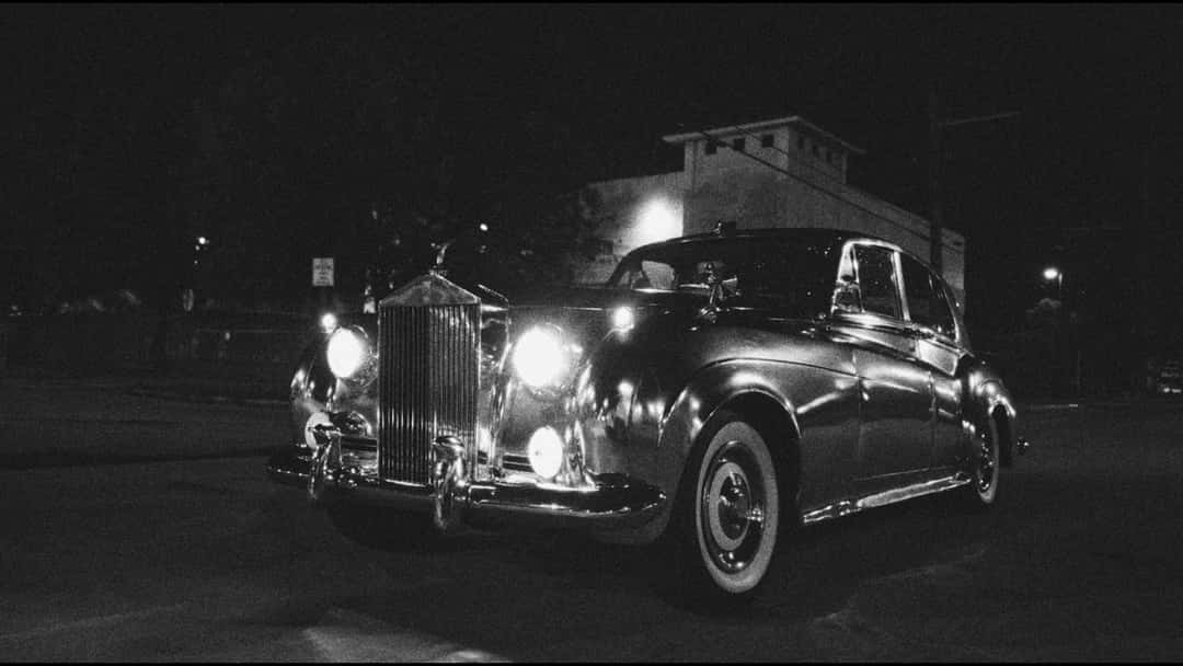 Un Rolls-Royce clásico con los faros encendidos, de noche frente a un edificio, en blanco y negro, en una imagen del video de Basic Burner
