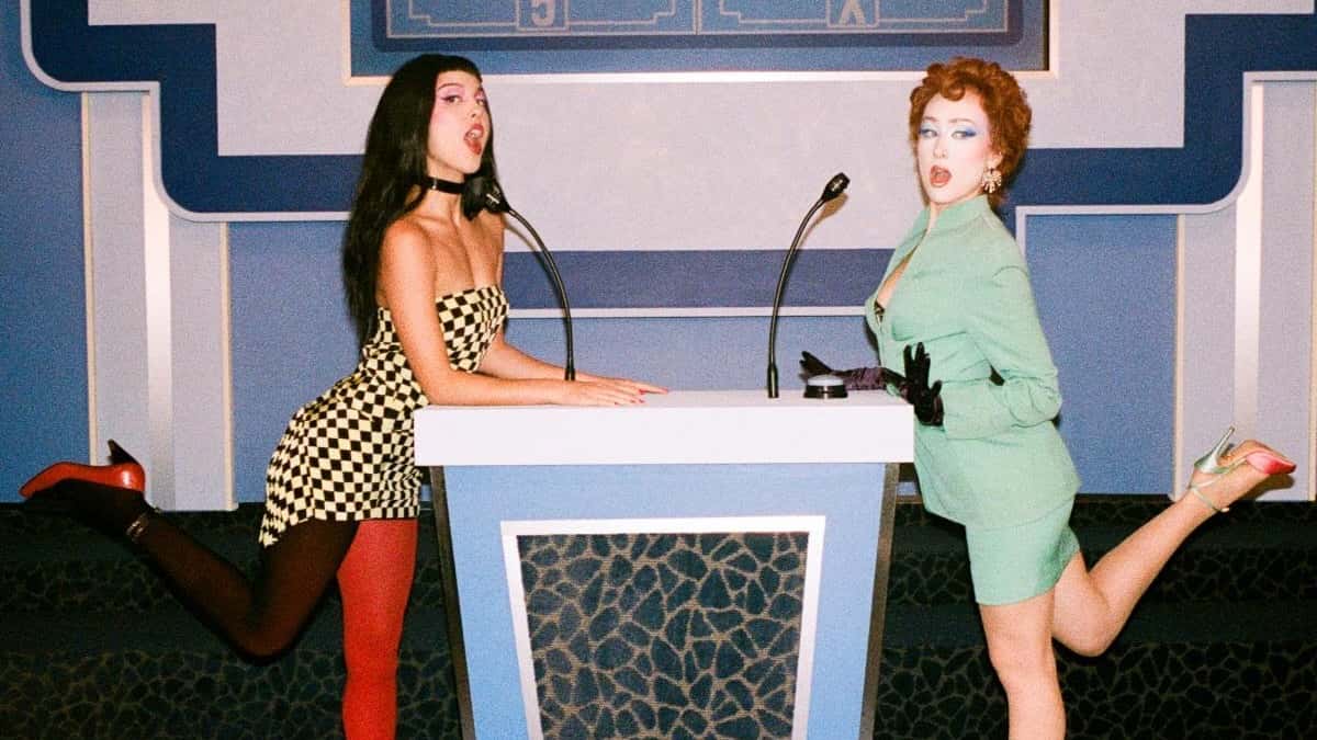 Foto de prensa oficial del remix de "expectations": Olivia Rodrigo, con vestido corto a cuadros blancos y negros, choker y una media roja y otra negra, y Chappell Roan, con traje verde agua, guantes negros y pelo rojo enrulado, posan con una pierna levantada a cada lado de un atril de concurso de TV con dos micrófonos, frente a un decorado azul. Foto: Phoenix Johnson