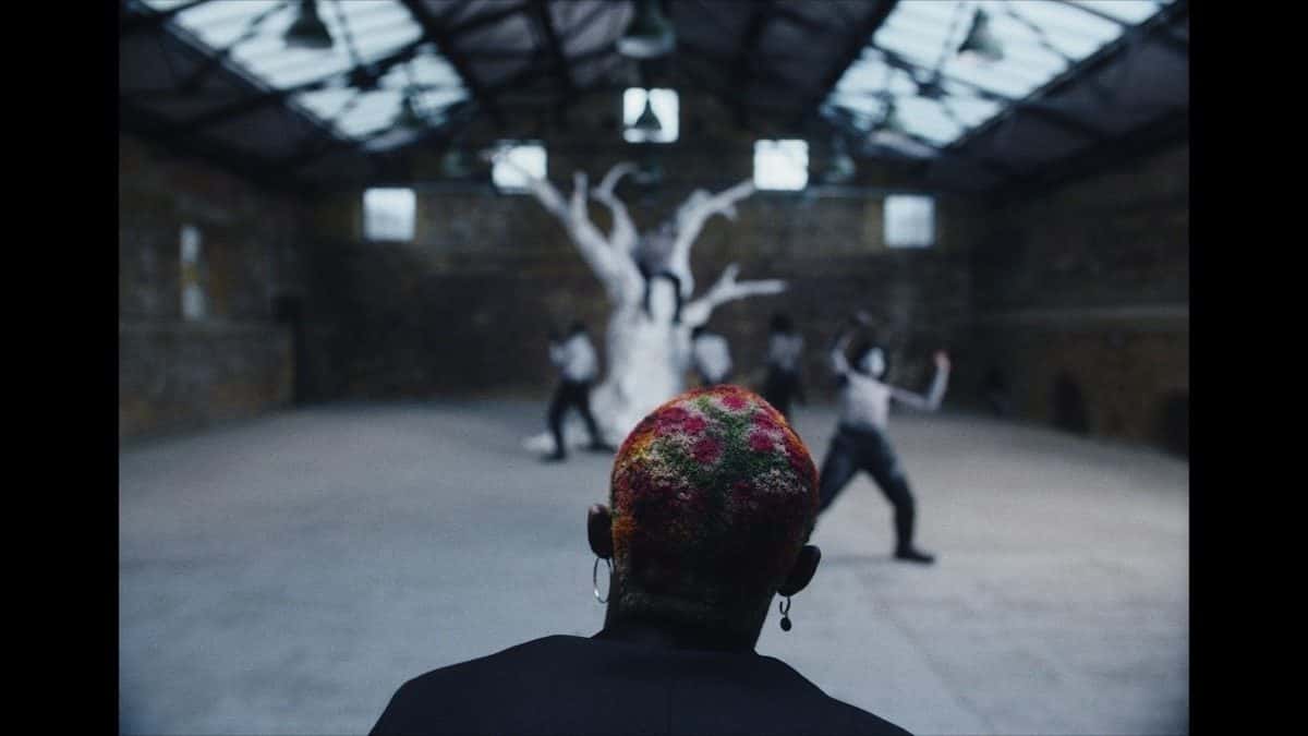 Omah Lay, de espaldas y con el pelo teñido de colores, mira a un grupo de bailarines alrededor de un árbol blanco dentro de un galpón con techo de vidrio, en una imagen de archivo del video oficial de «Artificial Happiness»