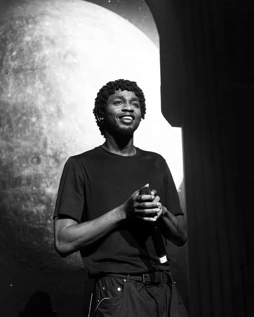 Foto de archivo de Omah Lay en 2023, en blanco y negro: sonríe de pie en un escenario, con remera negra y las manos juntas sobre el micrófono, frente a una pantalla con una luna gigante. Foto: Warner Records, Wikimedia Commons, CC BY-SA 4.0