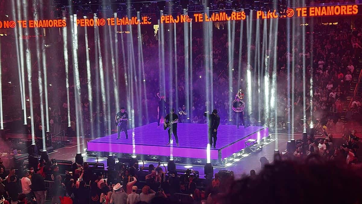 Foto de archivo de Fuerza Regida en vivo en el Capital One Arena de Washington, en octubre de 2024: la banda toca sobre una plataforma iluminada de violeta en el centro del estadio, rodeada de columnas de luz blanca y con el público alrededor. Foto: Floppykart, Wikimedia Commons, CC BY 2.0
