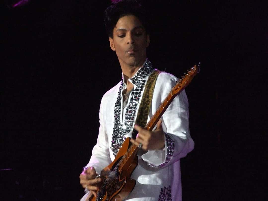 Prince en el festival Coachella de 2008, un año después del show de Las Vegas: toca una guitarra de madera con una camisa blanca bordada con pedrería, sobre un fondo oscuro (foto de archivo). Foto: penner, CC BY-SA 3.0, vía Wikimedia Commons