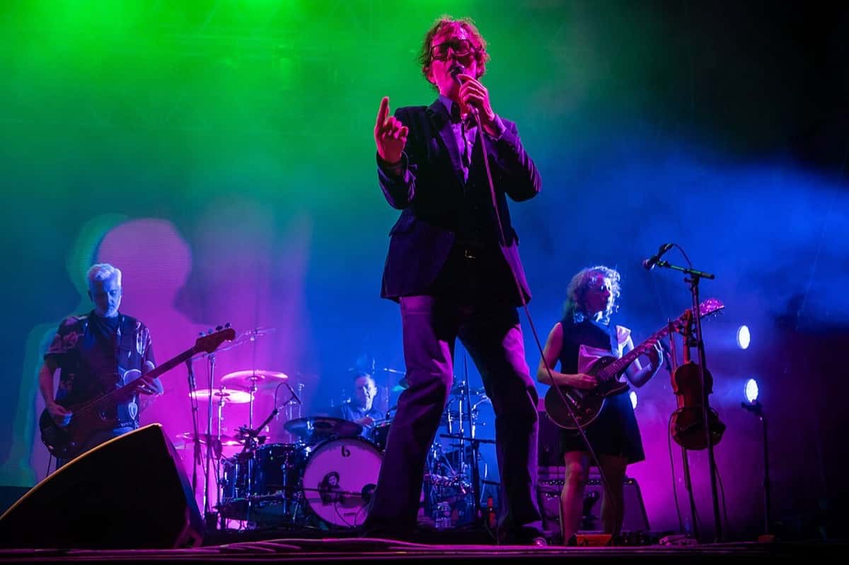 Jarvis Cocker canta con anteojos y traje oscuro en el centro del escenario, con el bajista a la izquierda, el baterista atrás y una guitarrista a la derecha, bajo luces verdes y violetas, en el festival La Route du Rock de 2025 (foto de archivo)
