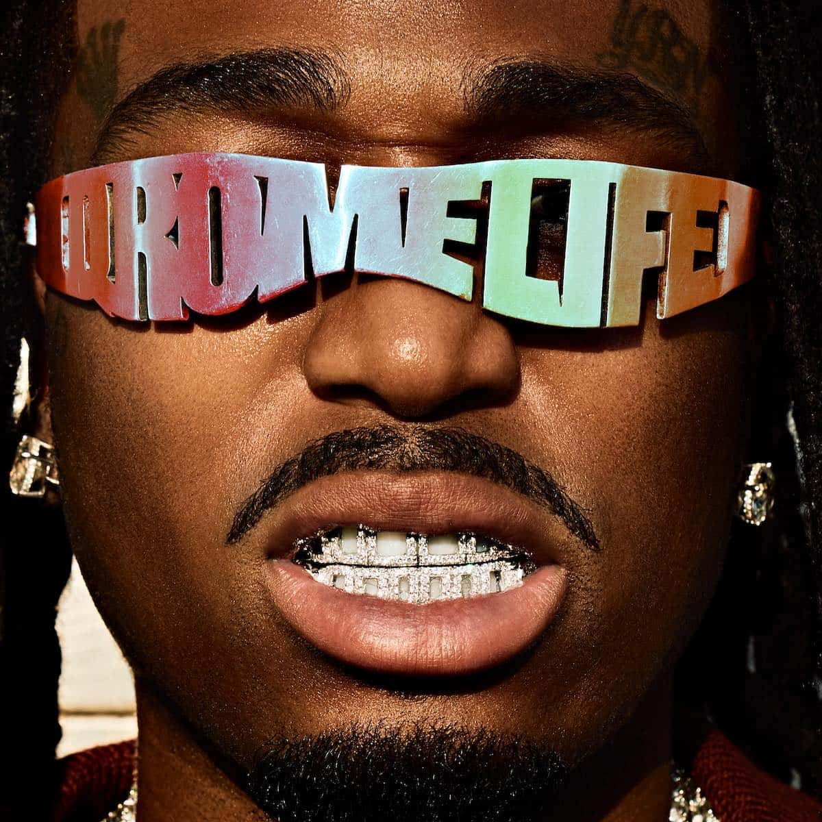 Primer plano de Quavo con unos anteojos metálicos que forman la palabra QRÖMELIFE y grillz de diamantes, imagen de su nuevo disco