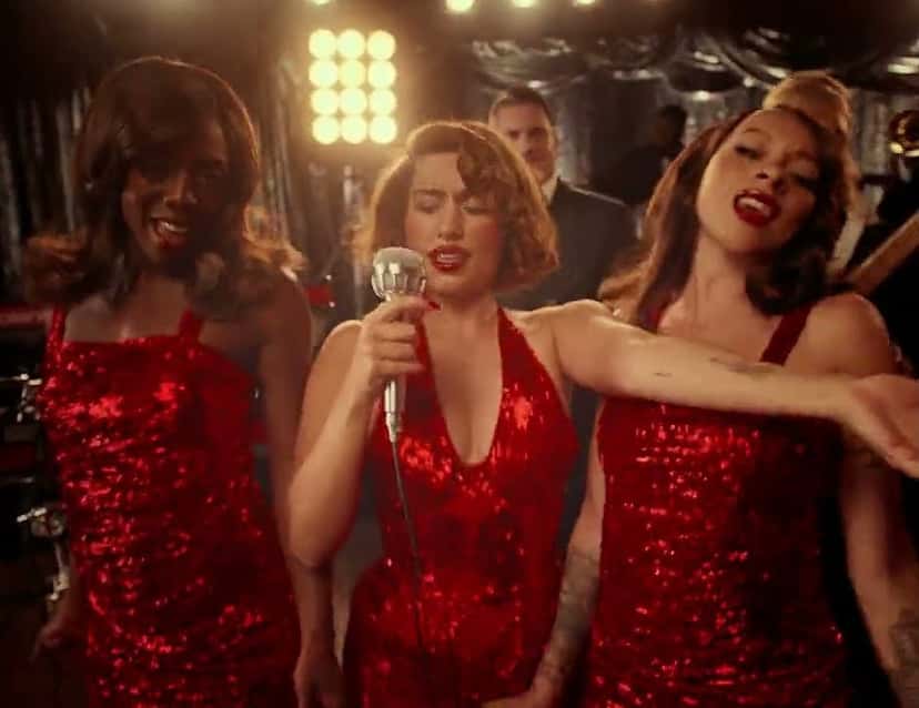 Fotograma del video oficial de «WHERE IS MY HUSBAND!»: Raye canta con un micrófono antiguo, con vestido rojo de lentejuelas, junto a dos coristas vestidas igual, en un escenario con luces (foto de archivo)