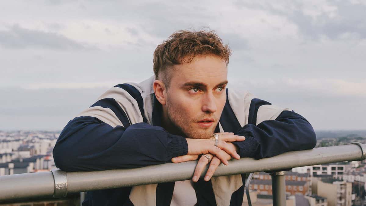 Foto oficial de Sam Fender: el músico apoyado con los brazos cruzados sobre una baranda en una terraza alta, con campera azul con mangas blancas, anillos en las manos y la ciudad de fondo bajo un cielo nublado