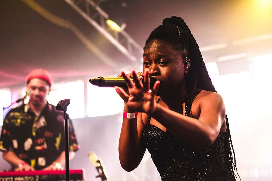 Sampa the Great canta con el micrófono tomado con las dos manos, con trenzas largas y vestido negro brillante, con un tecladista detrás, en el festival Haldern Pop de Alemania, en 2018 (foto de archivo). Foto: Martin Schumann, CC BY-SA 4.0, vía Wikimedia Commons
